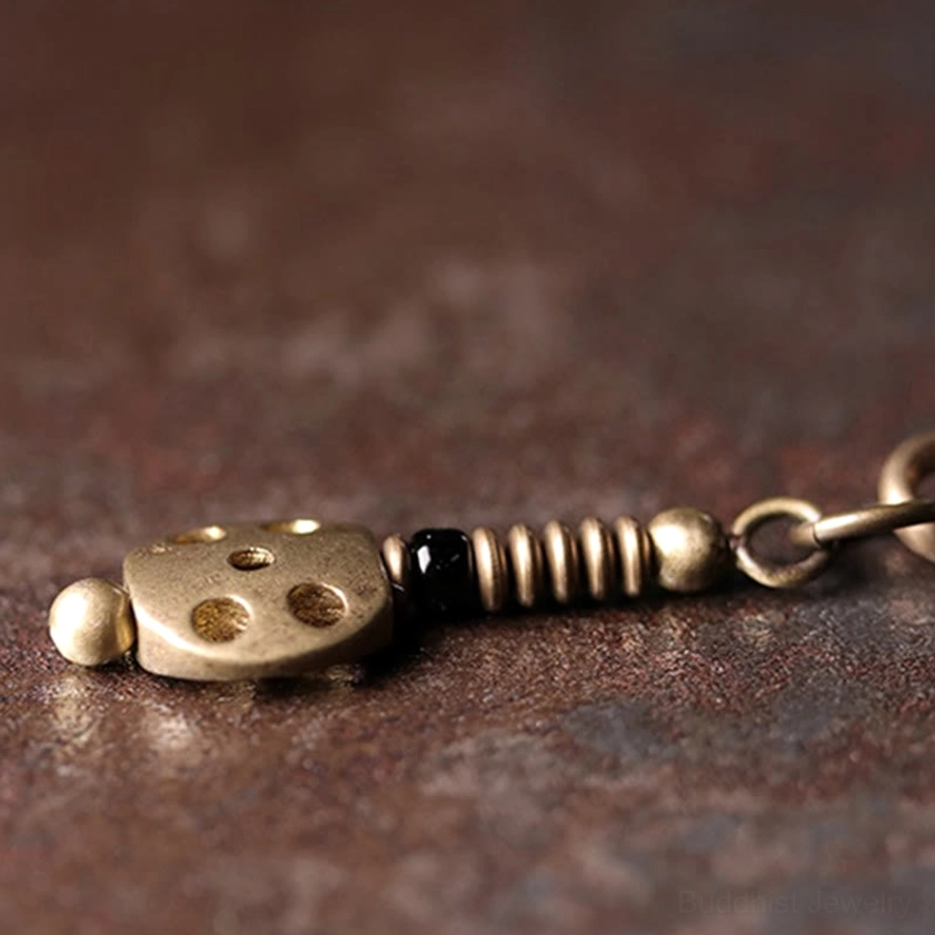 Macro close-up of brass dice-shaped tassel pendant with spring-style copper hardware and black beads on rainbow obsidian Buddhist bracelet