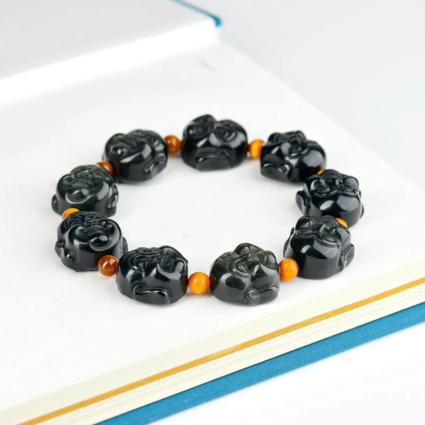 Top-down flat lay of rainbow obsidian Buddha bracelet on hardcover book, displaying carved beads and orange spacer details