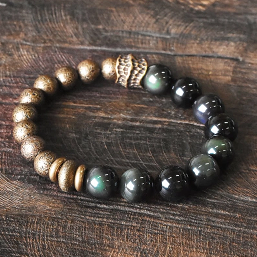 Oblique overhead view of rainbow obsidian and copper beaded bracelet with carved metal accents and round bronze spacers on wood