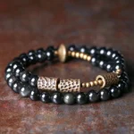 Rainbow obsidian double wrap bracelet with hammered copper accents, flat lay top view showing black glossy beads and bronze metal details on rustic tabletop