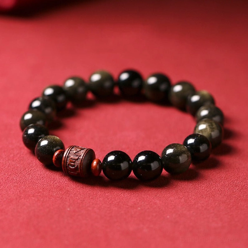 Rainbow Obsidian Om Mani Padme Hum bracelet with golden sheen beads and carved spiritual symbol bead on red backdrop