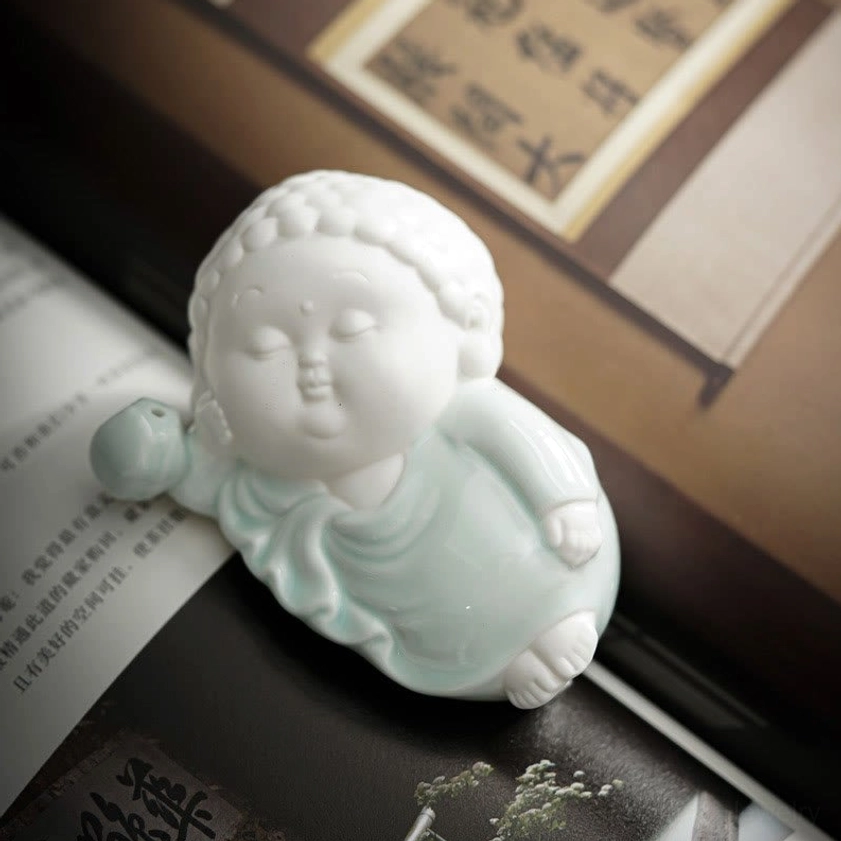 Side-lying baby Buddha ceramic ornament with celadon glazed robes - low-angle frontal shot bookside display