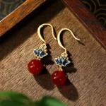 Red agate lotus drop earrings with blue enamel charm and gold-plated ear hooks, displayed on dark wooden tray in flat lay photography