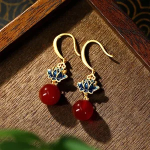 Red agate lotus drop earrings with blue enamel charm and gold-plated ear hooks, displayed on dark wooden tray in flat lay photography