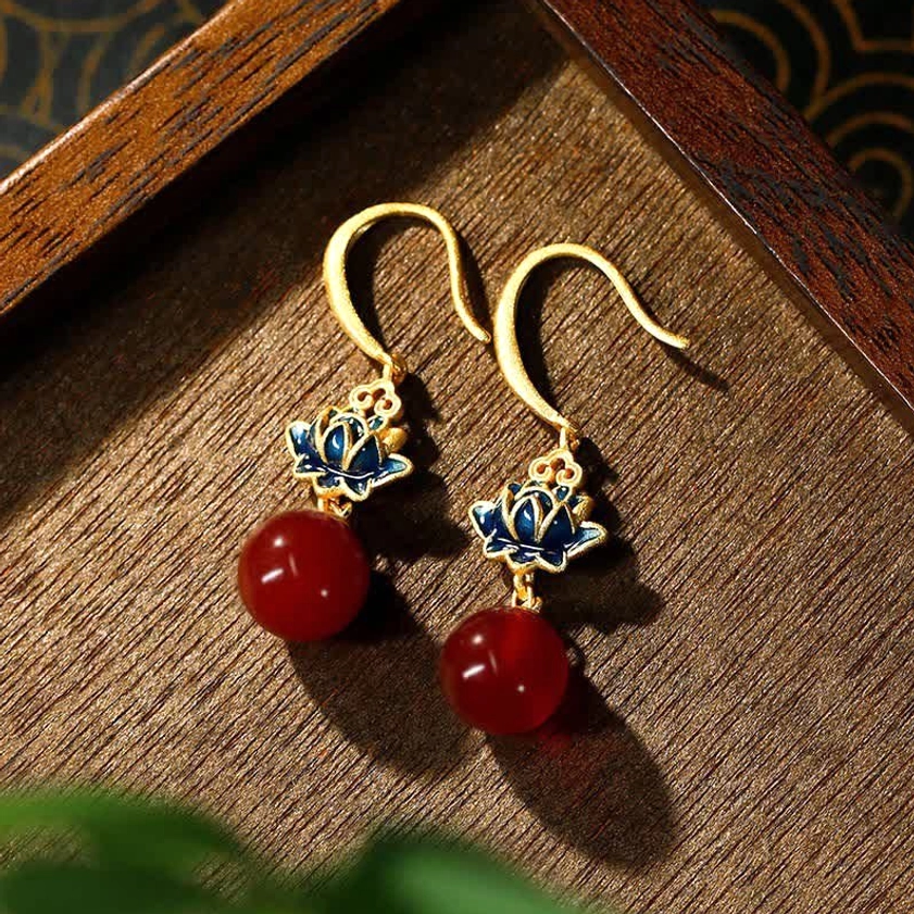 Red agate lotus drop earrings with blue enamel charm and gold-plated ear hooks, displayed on dark wooden tray in flat lay photography