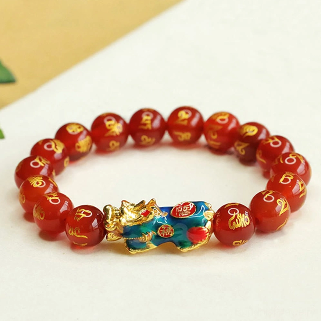 Angled top-down view of red agate Pixiu mantra bracelet on light paper, large red agate beads with prominent gold script, striking blue-gold Pixiu charm