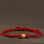 Red braided Buddhist string bracelet with gold Om Mani Padme Hum engraved bead, front view on dark background