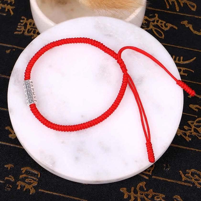 Single red Om Mani Padme Hum Buddhist protection bracelet with engraved silver charm on braided cord, displayed on marble plate with calligraphy background