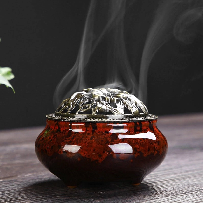Red ceramic Buddhist incense burner with bronze floral lid emitting fragrant smoke on wooden table