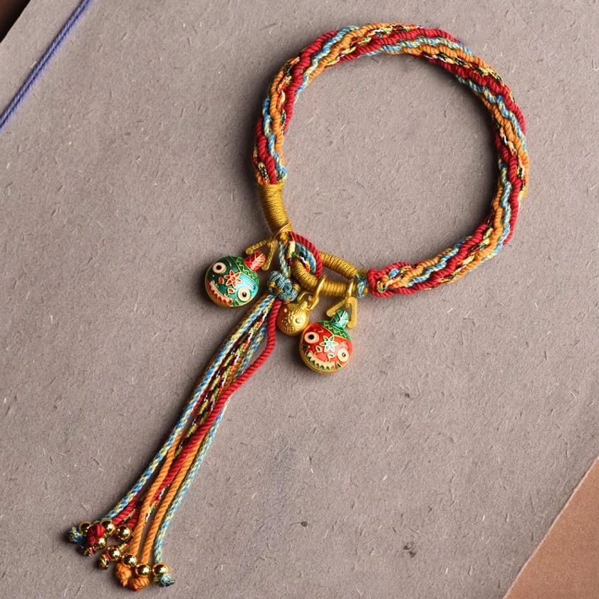Red and gold toned reincarnation knot braided string bracelet with enamel swallow charms and decorative tassel on gray background