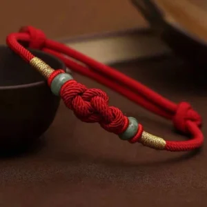Red string jade bracelet with gold accents on dark ceramic bowl, close-up showing knotted detail and green jade beads