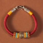 Red Buddhist King Kong Knot braided bracelet with engraved charm, turquoise and yellow beads, premium silver clasp on brown table