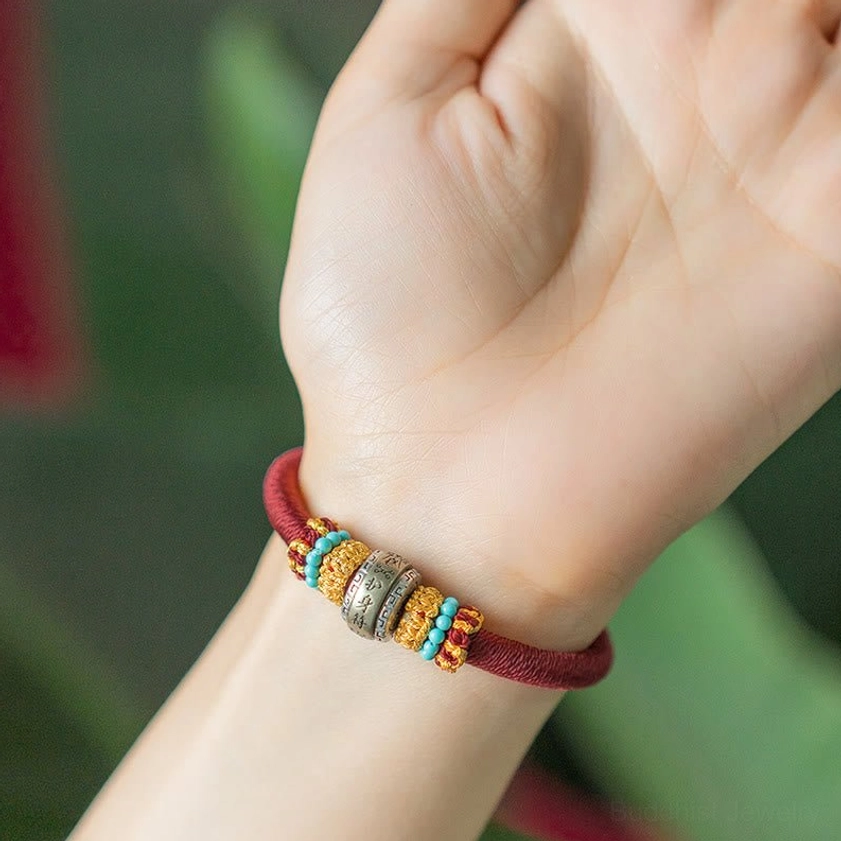 Red Buddhist lucky bracelet worn on wrist with green foliage background, showing King Kong Knot charm and bead details
