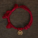 Red Buddhist lotus string bracelet flat lay showing burgundy braided cord with gold hollow lotus pendant and tasseled ends on wooden surface