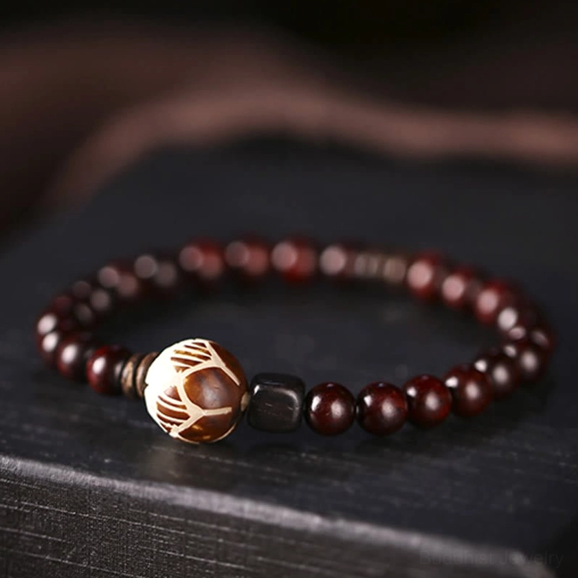 Close-up top-down view of single wrap wooden bracelet on dark wooden table, showing deep red sandalwood beads, hand-carved lotus focal bead, warm lighting