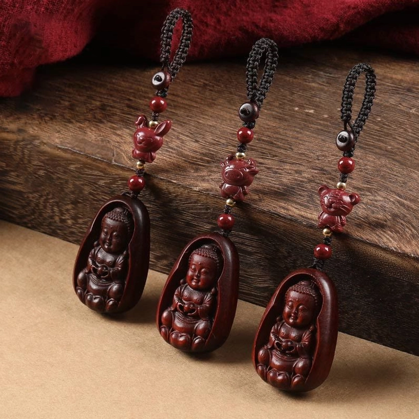 Red sandalwood Buddha zodiac keychains flat lay with black braided cord and red cinnabar beads on wooden table with fabric backdrop