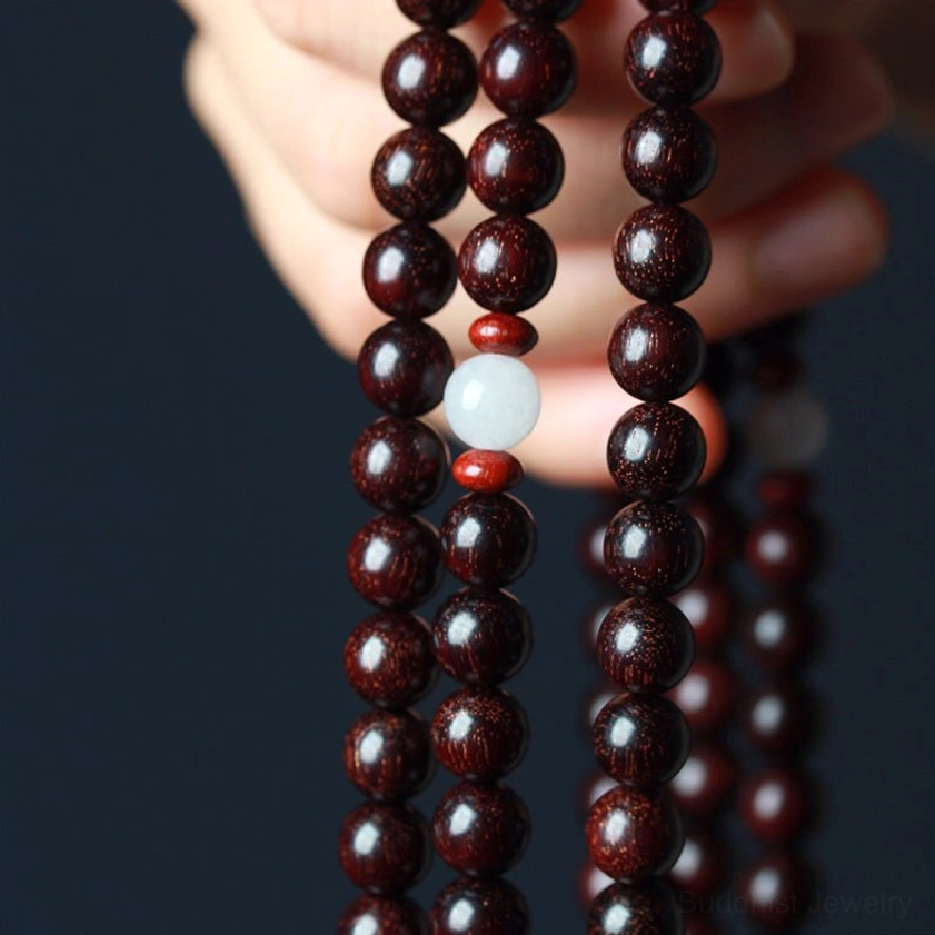 Detailed front view of two hands holding sandalwood prayer mala displaying glossy reddish-brown wood grain white and red accent beads