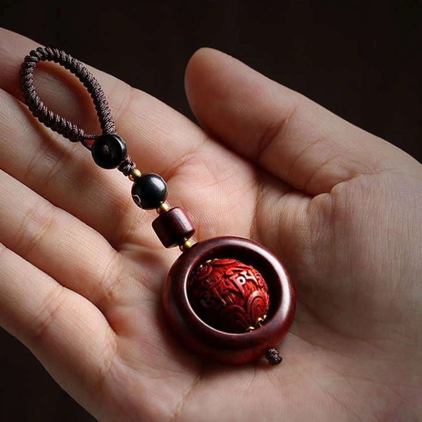 Single red sandalwood prayer keychain pendant held in human hand with carved central sphere and braided rope lanyard