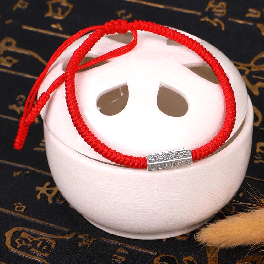 Red Buddhist string bracelet with vintage silver ornament and knotted braided rope design shown next to white ceramic jar with calligraphy backdrop, adjustable drawstring