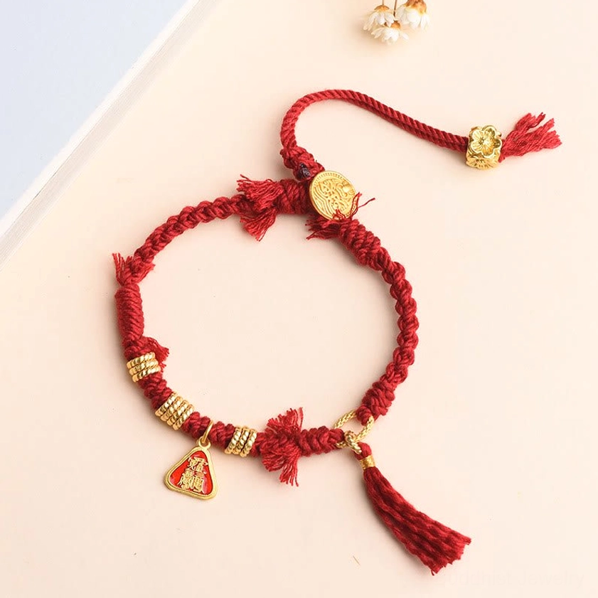 Buddhist protection bracelet featuring red braided rope, golden goddess charm, and decorative flower tassel on light beige background