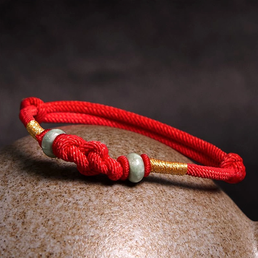 Red string couple bracelet with central braided knot, gold-wrapped cord section, and jade bead decoration on ceramic surface