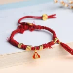 Red string protection bracelet with goddess charm and flower tassel, displayed flat on book edge with braided cord and gold metal accents