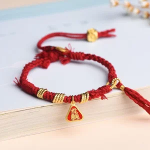 Red string protection bracelet with goddess charm and flower tassel, displayed flat on book edge with braided cord and gold metal accents