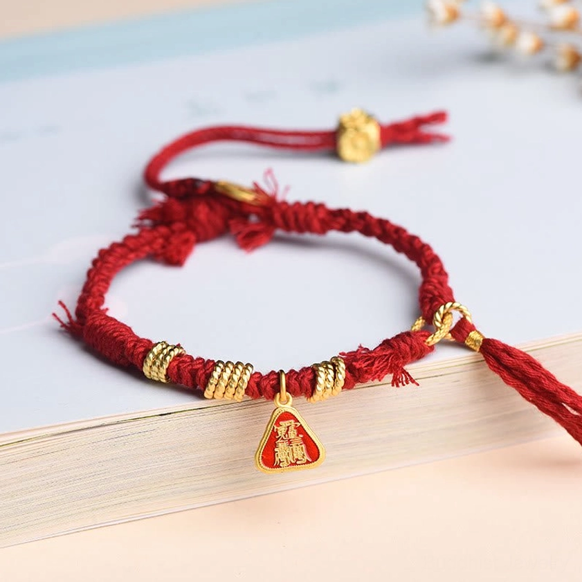 Red string protection bracelet with goddess charm and flower tassel, displayed flat on book edge with braided cord and gold metal accents
