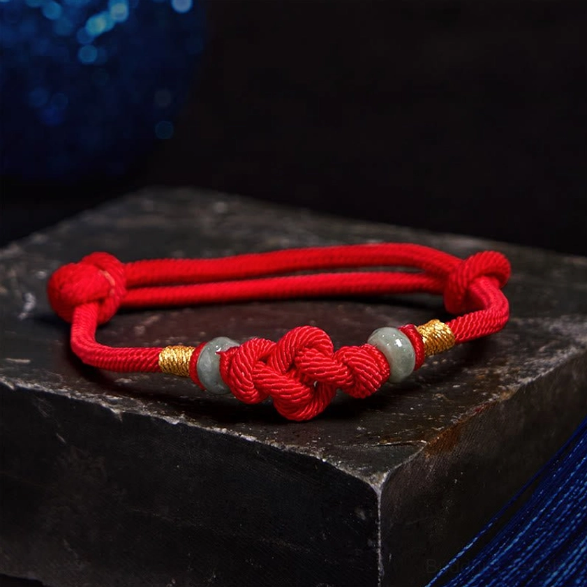 Lucky red string bracelet with braided knot, gold metal accents, and jade stone beads on black stone surface