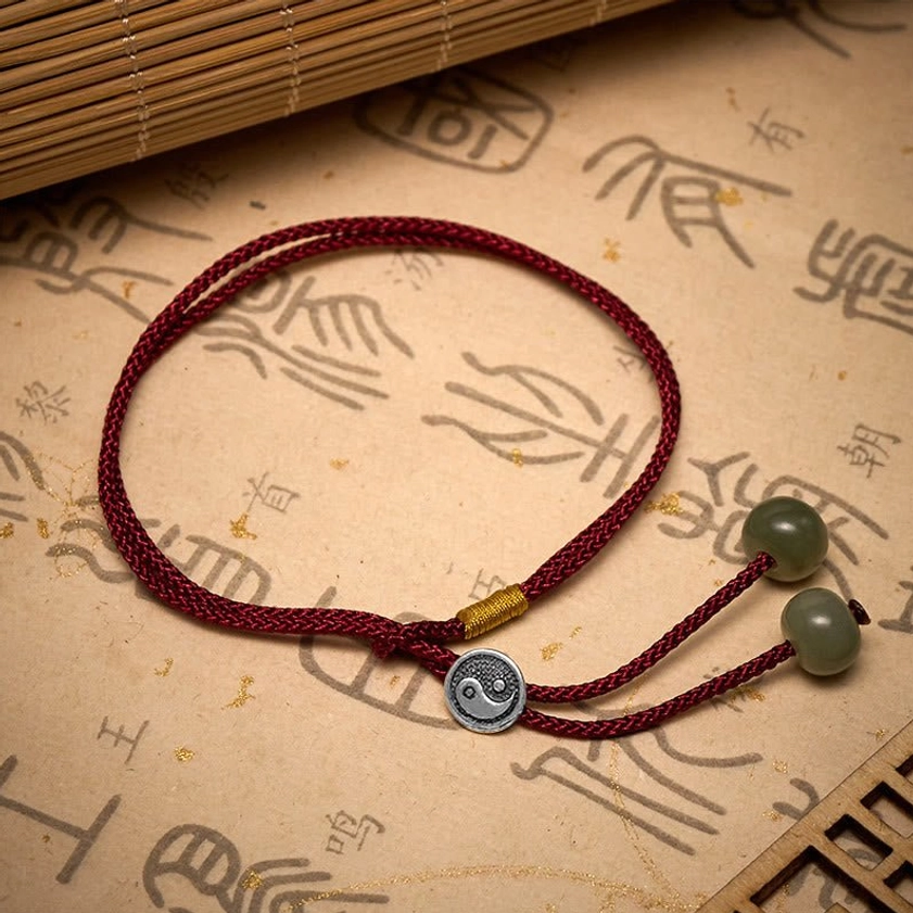 Flat lay of Yin Yang Buddhist red string bracelet with braided cord, silver Yin Yang charm, green stone beads, and adjustable tassel on bamboo mat