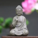 Serene resin Buddha statue with Abhaya hand mudra in matte light gray, seated in lotus position on wooden table with pink flower background - meditation desk decoration