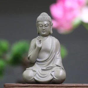 Serene resin Buddha statue with Abhaya hand mudra in matte light gray, seated in lotus position on wooden table with pink flower background - meditation desk decoration