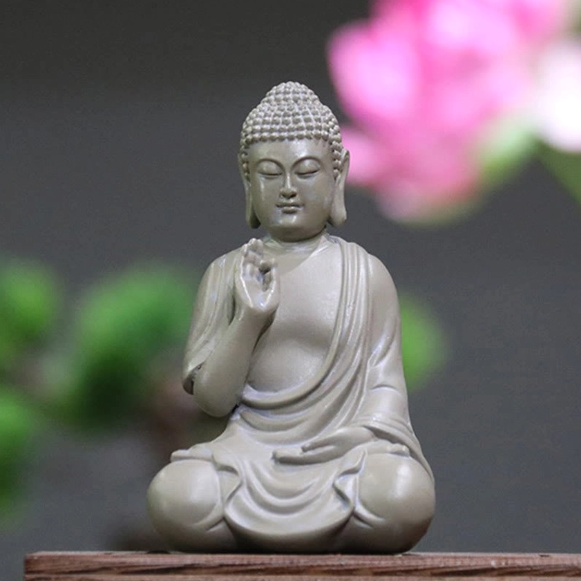 Serene resin Buddha statue with Abhaya hand mudra in matte light gray, seated in lotus position on wooden table with pink flower background - meditation desk decoration