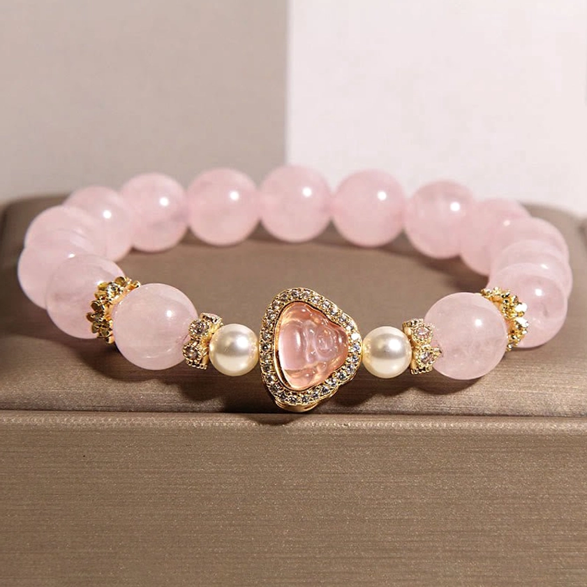 Rose quartz Buddha bracelet with gold rhinestone accents displayed on brown jewelry tray, side overhead view showing soft pink crystal beads and decorative Buddha charm