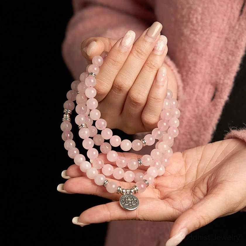 Two hands holding rose quartz 108 mala beads with silver lotus charm, wearing meditation bracelet