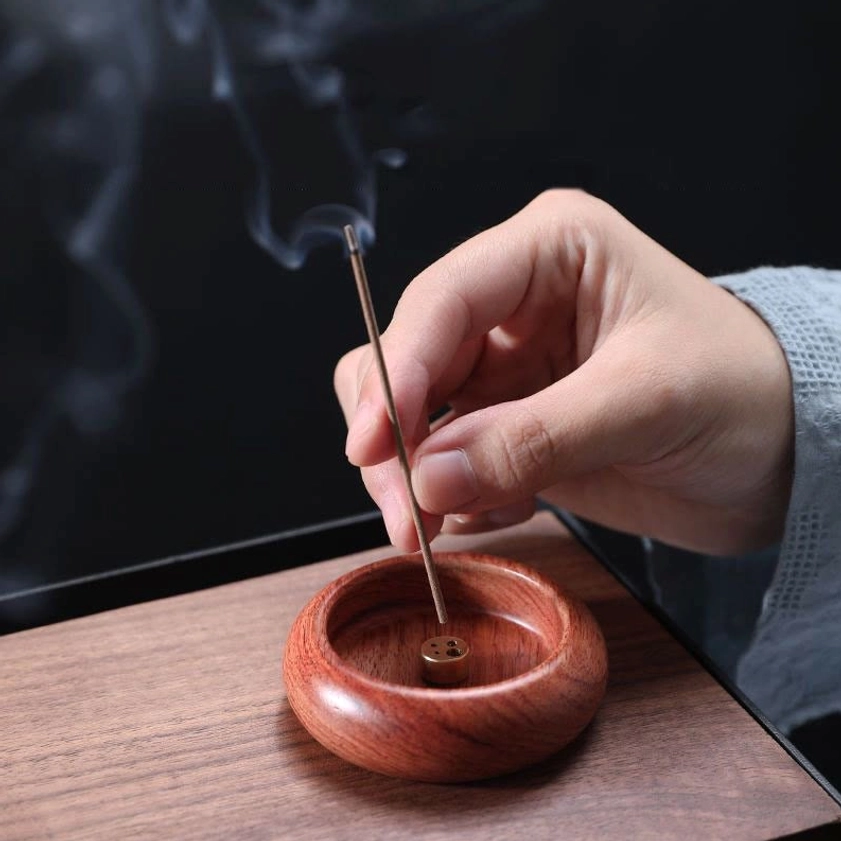 Light brown rosewood incense burner on wooden table with hand inserting smoking incense stick