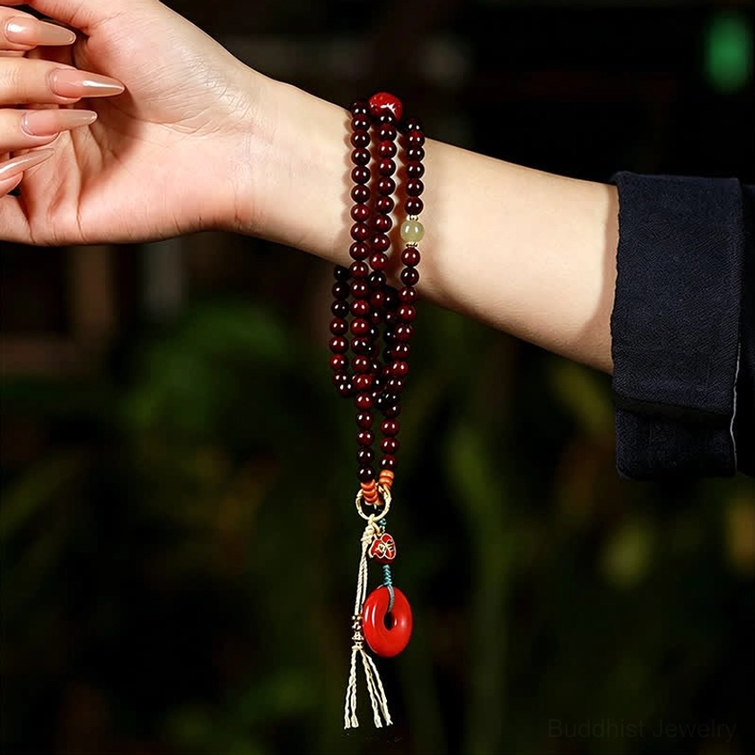Red rosewood 108 bead mala worn on human wrist in close-up side view with green foliage background, showing deep red beads and peace buckle pendant