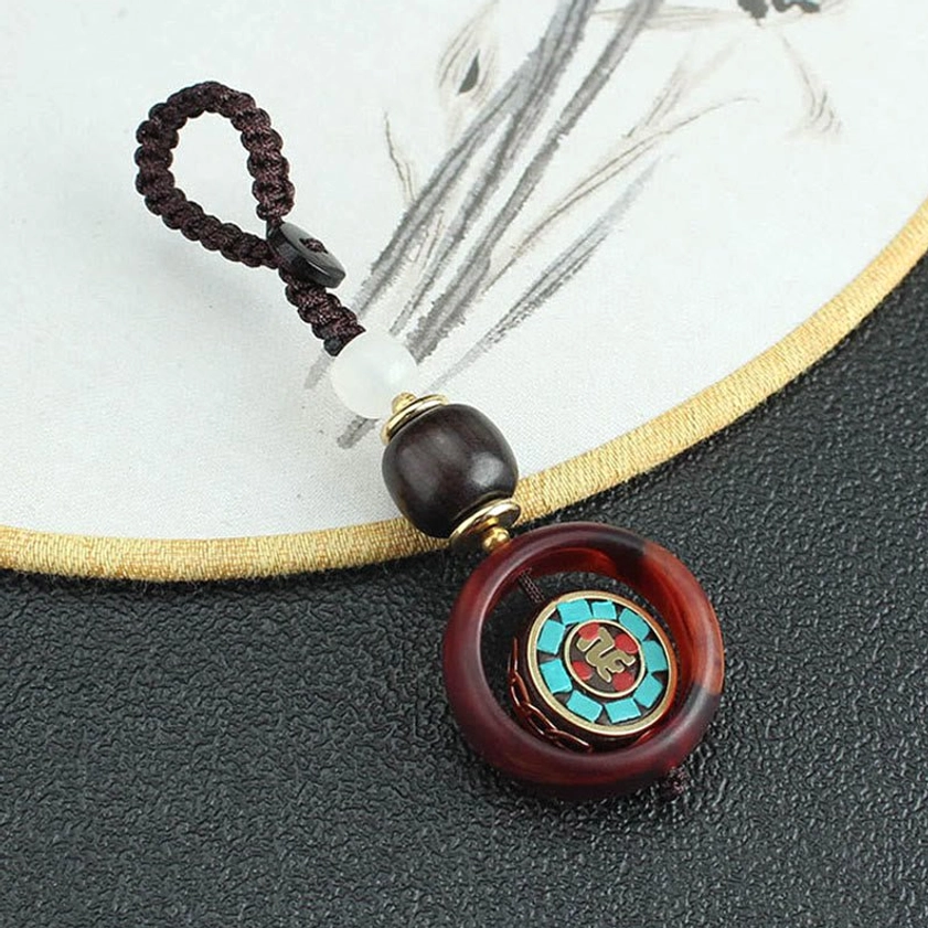 Overhead view of rotating Sanskrit Buddhist charm keychain on leather mat with Chinese fan, showing braided brown cord and wooden beads