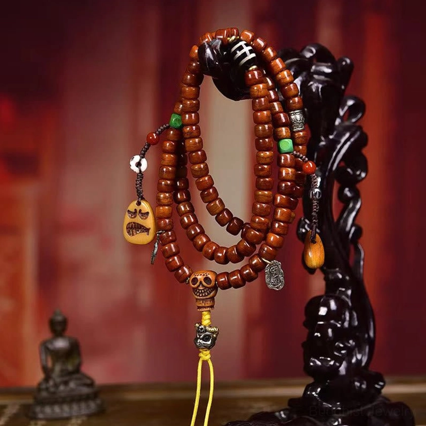 Buddhist prayer mala bracelet hanging on black wood stand with small Buddha statue background, showing reddish-brown beads and metal decorations