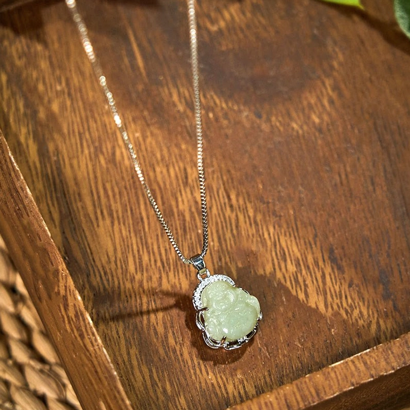 Light green Hetian jade Laughing Buddha Buddhist pendant with silver bezel and sparkling crystal halo, shot from oblique angle on dark wooden surface - prosperity and abundance necklace