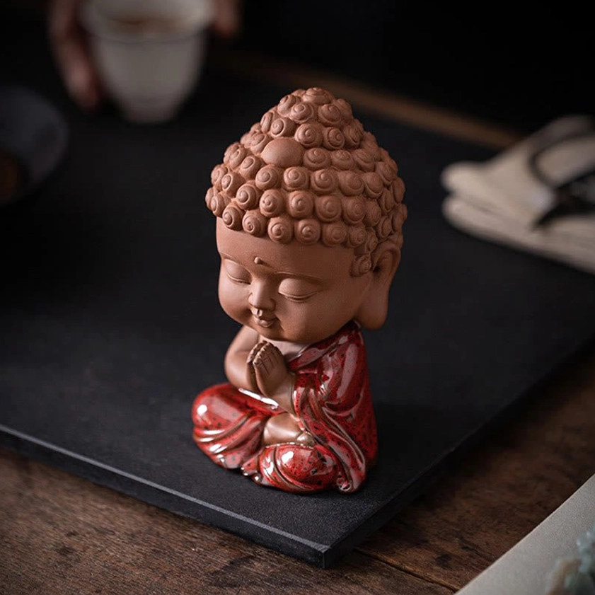 Side three-quarter view of mini Sakyamuni Buddha in cross-legged sitting pose with closed eyes, red glazed robes, displayed on dark mat and wooden table