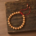Sandalwood mala bracelet with tan beads and red braided cord on dark wooden surface, top-down view