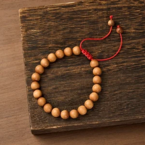 Sandalwood mala bracelet with tan beads and red braided cord on dark wooden surface, top-down view
