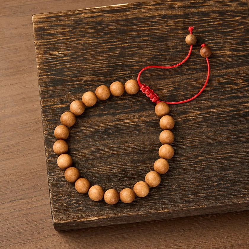 Sandalwood mala bracelet with tan beads and red braided cord on dark wooden surface, top-down view