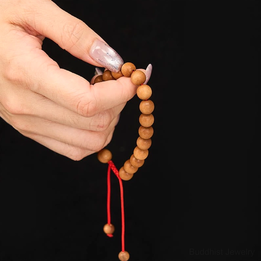 Close-up hand-held view of sandalwood mala bracelet with tan wooden beads and red adjustable cord held by fingers