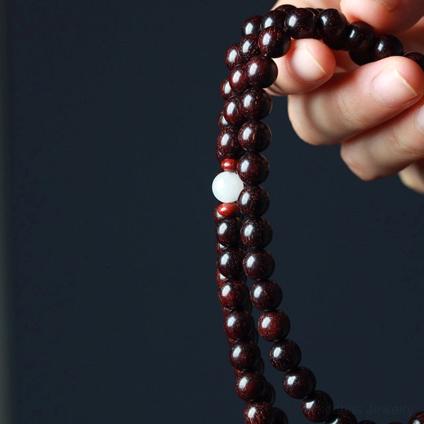 Finger-held close-up perspective of red sandalwood 108-bead mala showing polished beads white and red spacer beads on dark background