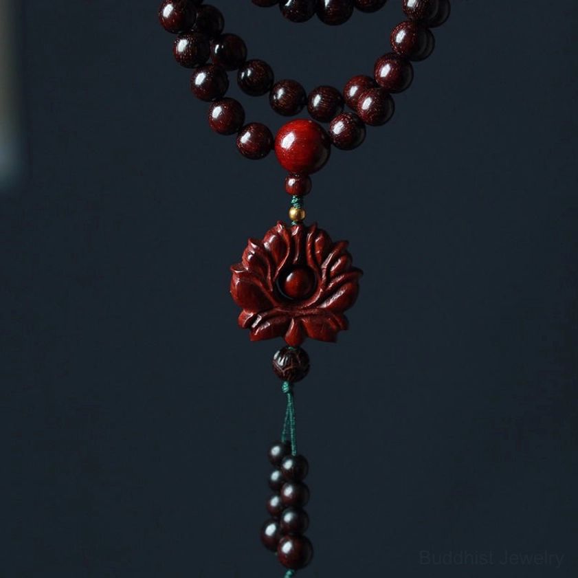 Flat lay display of red sandalwood 108-bead mala with central accent bead carved lotus pendant and green tassel on dark background