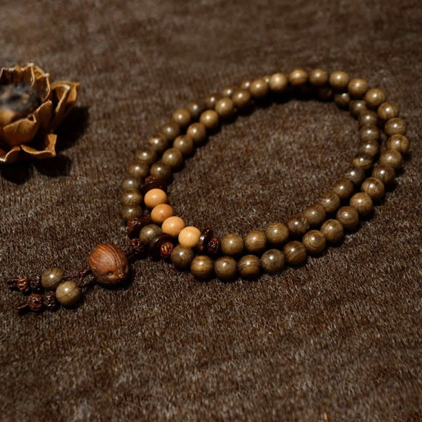 Single-loop sandalwood mala prayer beads with gradient brown wooden beads and decorative tassel, side view on fabric with botanical accents