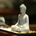 Handcrafted sandstone sitting Buddha statue in zen garden with gravel tray, left three-quarter view showing intricate robe details and meditation pose
