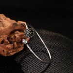 Silver lotus open cuff bangle with detailed bloom and bud charm, left side view on natural driftwood background